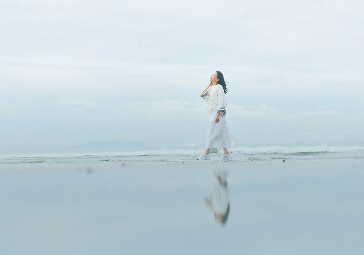 画像：砂浜を白い服を着た女性がリラックスして歩いている様子